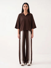 A model stands against a plain white studio background wearing a dark brown short-sleeve collared top and matching wide-leg trousers with vertical cream stripes.