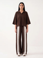A model stands against a plain white studio background wearing a dark brown short-sleeve collared top and matching wide-leg trousers with vertical cream stripes.