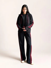 Model wearing Coastline’s black tracksuit with her hands in the hoodie pockets, showing the red sleeve stripes and coordinating trouser stripes.