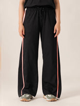Walking view of the Ellioti black flared trousers showing contrast piping along the legs and flowing hems over sneakers.