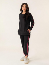Model wearing the Ivy black summer athleisure set with a long top and matching trousers with contrasting binding, with one foot forward.
