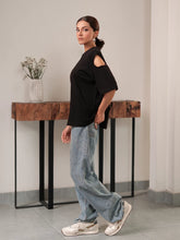 Model wearing Coastline's Lia Top in Black, showing the distinctive shoulder cut-out and relaxed boxy fit, styled with blue wide-leg jeans.