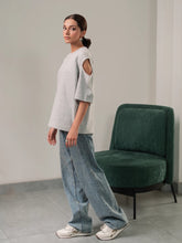 Model wearing Coastline's Lia Top in Heather Grey, showing its distinctive shoulder cut-out and relaxed boxy fit, styled with blue wide-leg jeans.