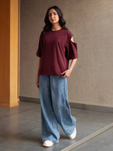 Model wearing Coastline's Lia Top in Mulberry, showing its relaxed boxy silhouette and shoulder cut-out, styled with light blue wide-leg jeans.
