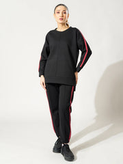 Front view of the Ora black co-ord set showing the loose drop-shoulder top and matching trousers, with one foot crossed in front.