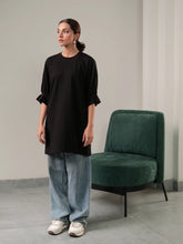 Model wearing Coastline's Tia Top in Black, featuring pleated sleeves and a relaxed longline silhouette, styled with light-wash wide-leg jeans.