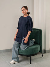 Model wearing Coastline's Tia Top in Oxford Blue, showing its relaxed longline fit and pleated sleeve detailing while seated on a green chair.