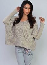 A female model wearing a beige chunky sweater with long sleeves and a round neckline, paired with gray pants. The model is posing with one hand behind her head.