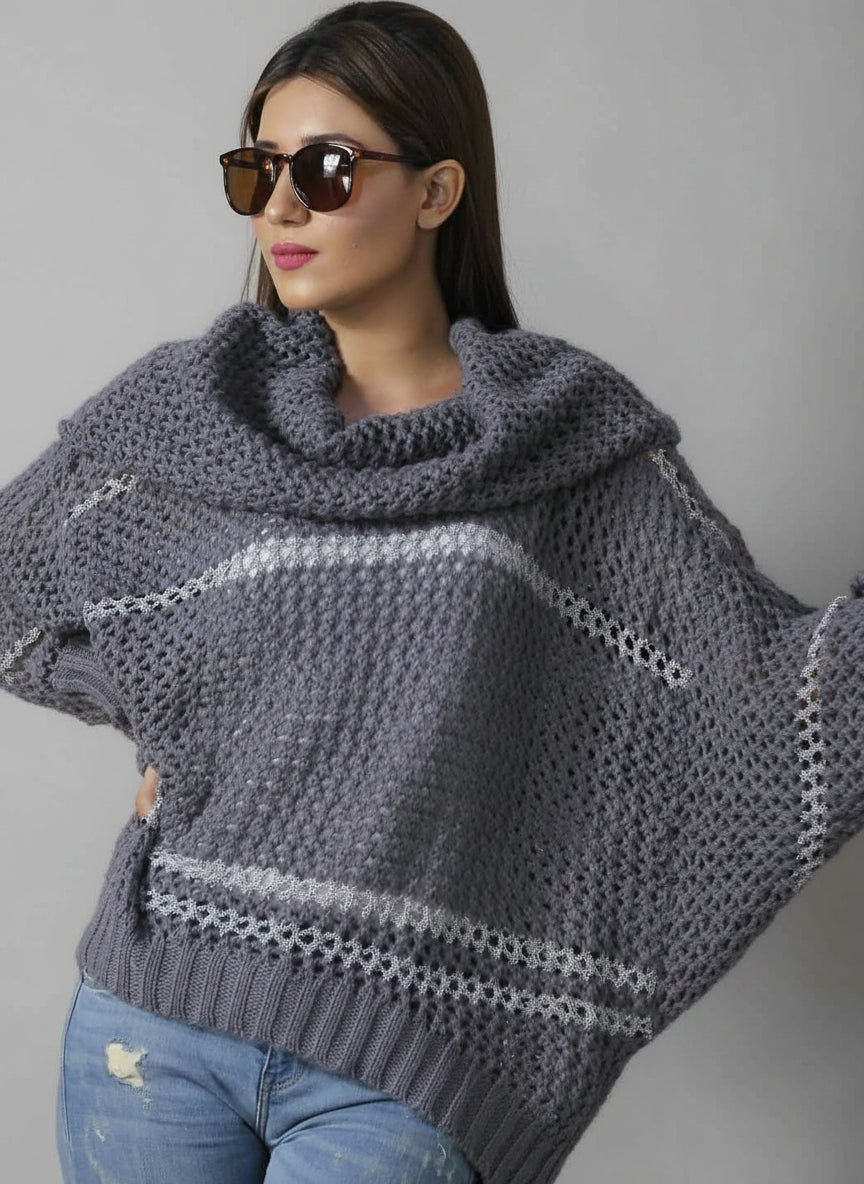 A woman wearing sunglasses and a gray turtleneck chunky sweater with batwing sleeves, standing in front of a newspaper print background.