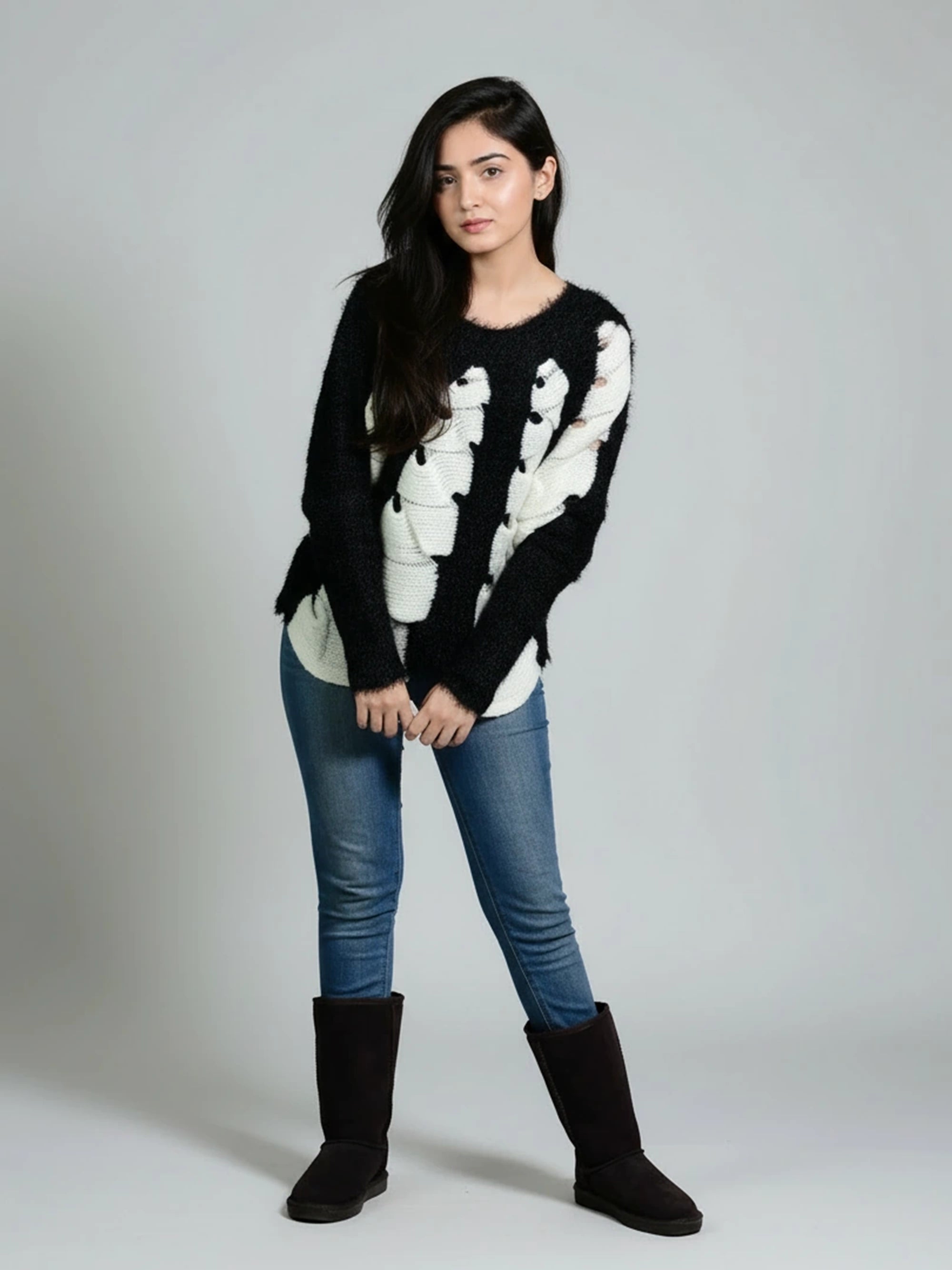 Woman wearing a black sweater with white patterns, blue jeans, and black boots on a gray background