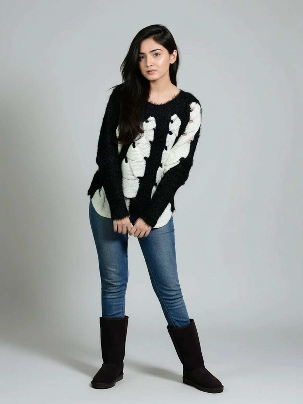 Woman wearing a black sweater with white patterns, blue jeans, and black boots on a gray background