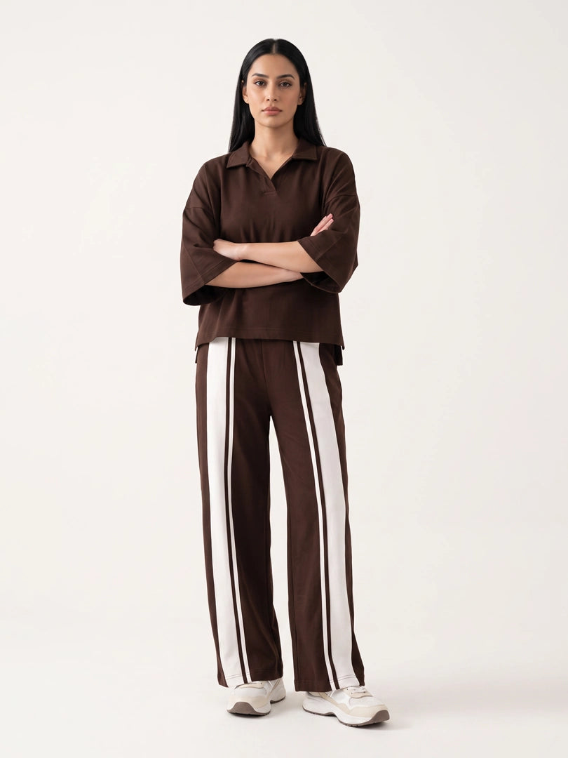 A woman stands against a plain light background wearing a dark brown collared short-sleeve top and matching wide-leg pants with bold white side stripes, paired with white sneakers.