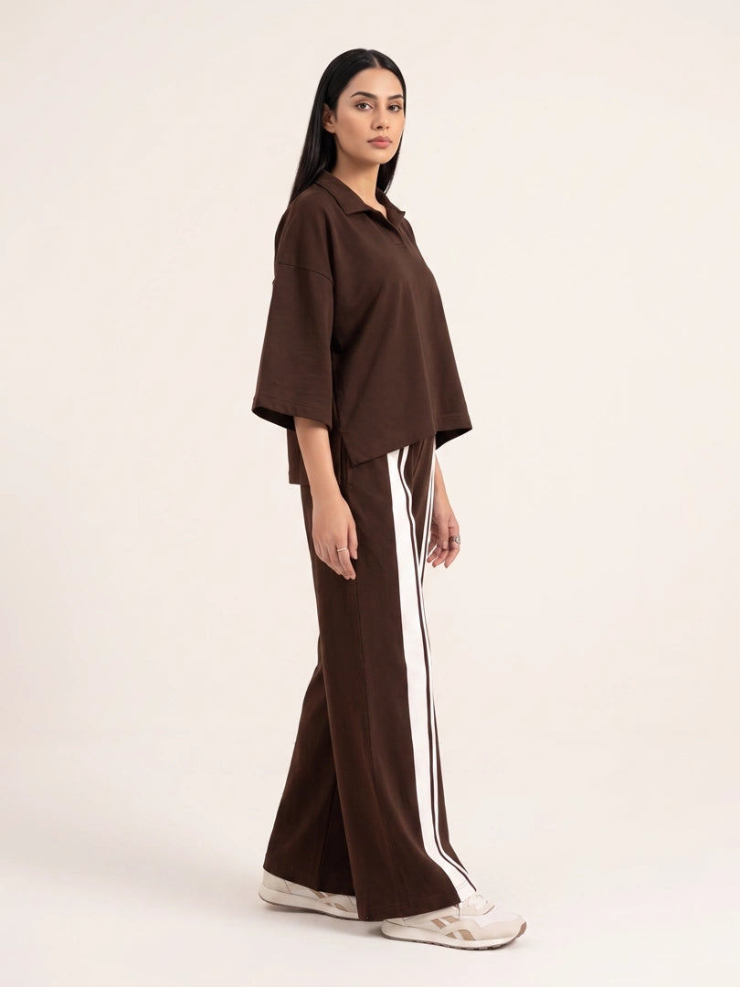 A woman modeling a dark brown two-piece outfit consisting of a short-sleeve collared top and matching wide-leg pants with a white side stripe, set against a plain off-white studio background.