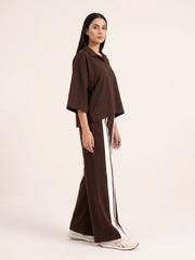 A woman modeling a dark brown two-piece outfit consisting of a short-sleeve collared top and matching wide-leg pants with a white side stripe, set against a plain off-white studio background.