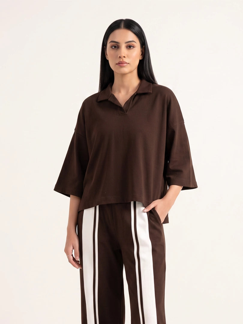 A woman modeling an oversized dark brown short-sleeve collared top with matching brown pants featuring white side stripes, against a plain off-white studio background.
