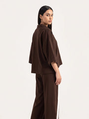 A woman modeling a matching dark brown lounge set consisting of an oversized short-sleeve top and wide-leg trousers, shown from a side-back angle against a plain white studio background.
