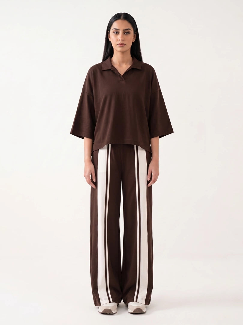 A model stands against a plain white studio background wearing a dark brown short-sleeve collared top and matching wide-leg trousers with vertical cream stripes.