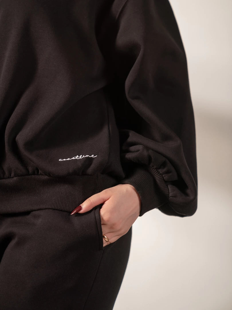 Detail view of the Barlow black fleece sweatshirt showing its balloon sleeve and ribbed hem, styled with black trousers.