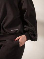 Detail view of the Barlow black fleece sweatshirt showing its balloon sleeve and ribbed hem, styled with black trousers.