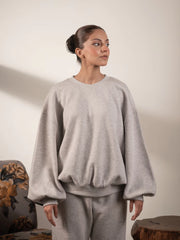 Model wearing the Barlow balloon sleeve sweatshirt in Heather Grey with her hands in her trouser pockets, showing its loose fit and gathered hem.