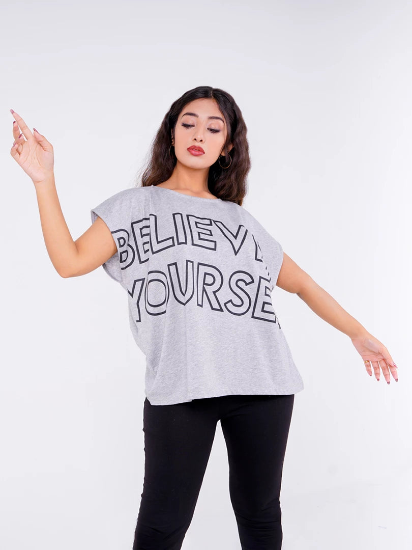 Model posing playfully with raised arms in the Believe In Yourself graphic top in Ash Grey, showing its loose boxy cut and oversized lettering.