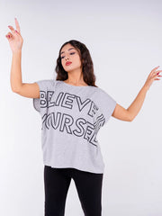 Model raising both arms while wearing Coastline's Believe In Yourself Ash Grey statement top, highlighting its bold front graphic and relaxed shape.
