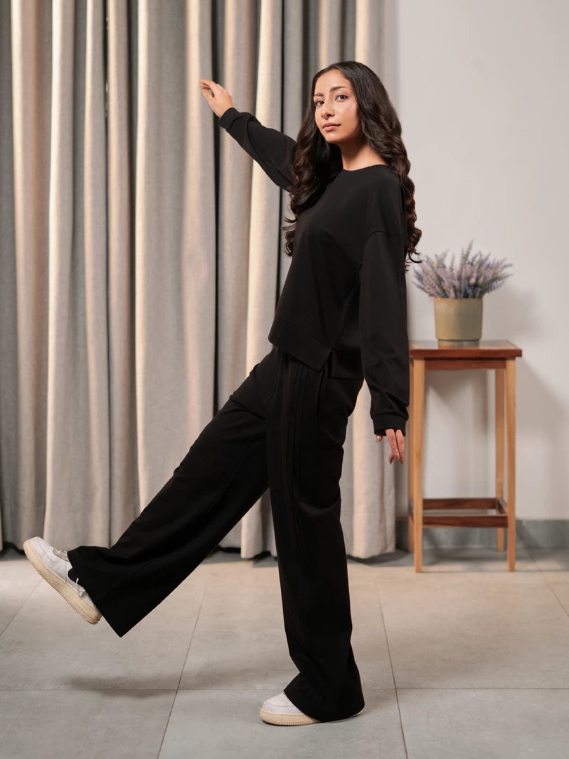 Model wearing the Brooklyn black summer co-ord set with a boxy long-sleeve top and wide-leg trousers, with one leg raised.
