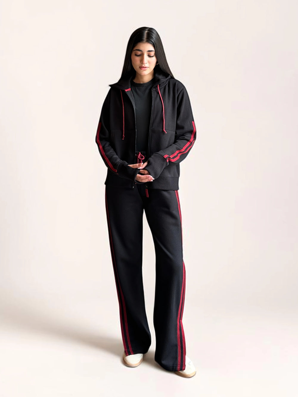 Model wearing Coastline’s black tracksuit with her hands in the hoodie pockets, showing the red sleeve stripes and coordinating trouser stripes.