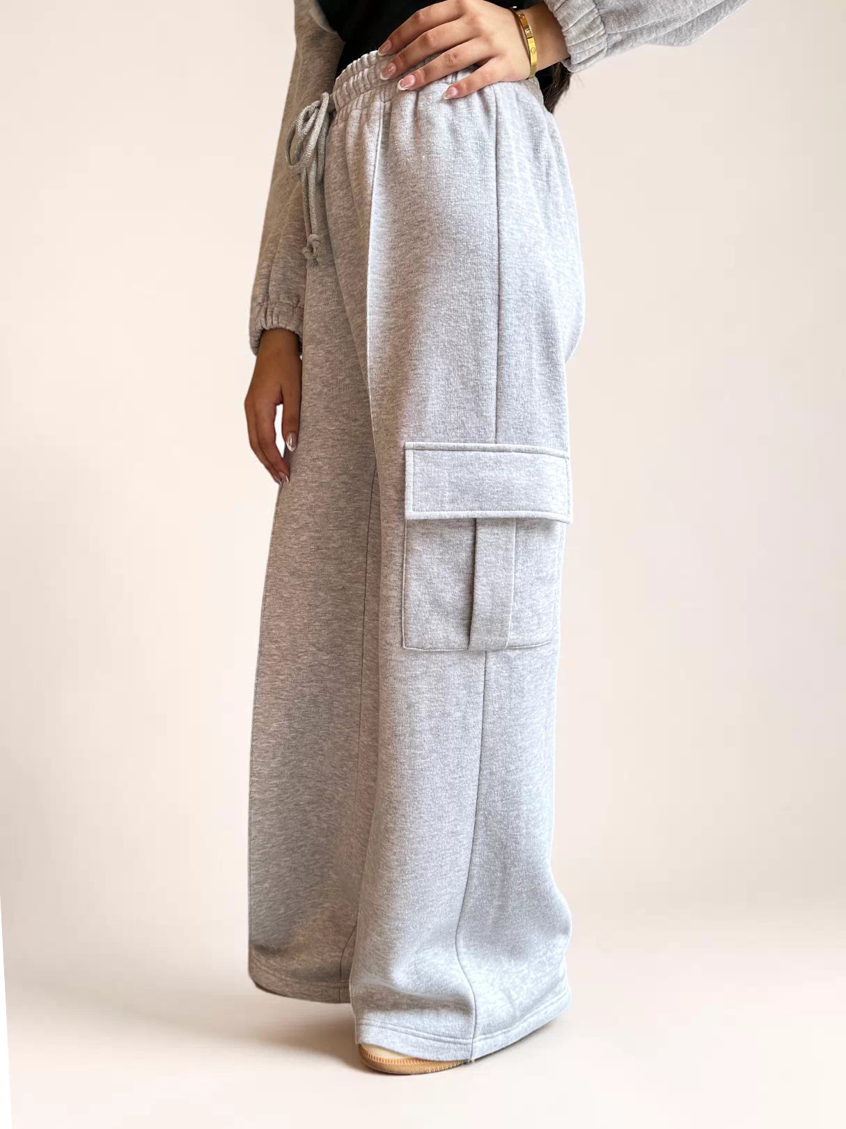 Side view of Coastline’s heather gray wide-leg cargo trousers, showing the flap thigh pocket and fleece fabric draping over cream shoes.