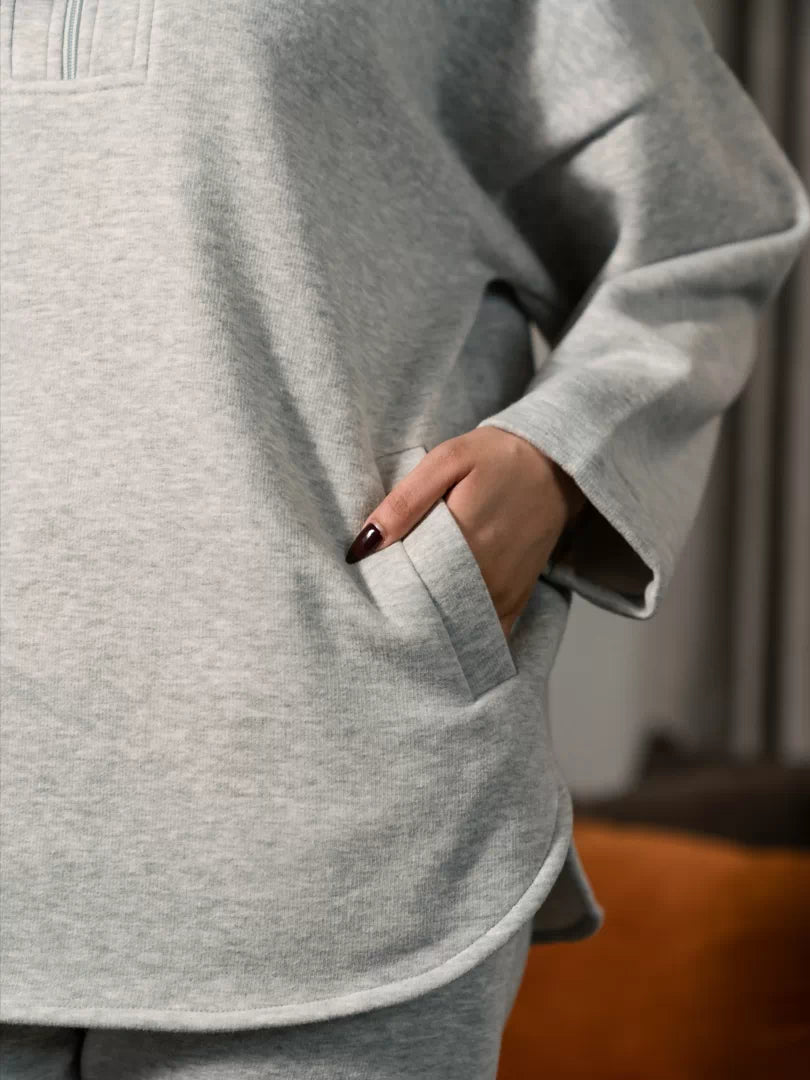 Close-up of the Darya Heather Grey hoodie showing a front welt pocket and curved hem, with one hand in the pocket.