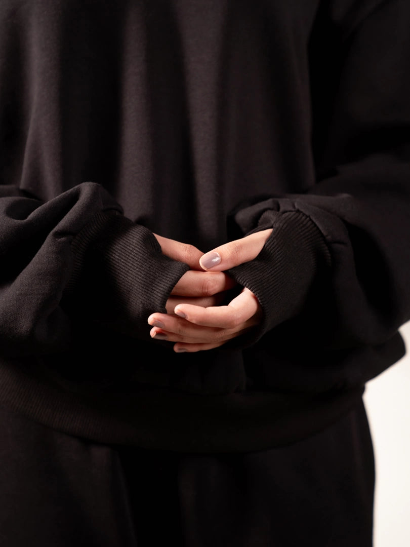 Front close-up of the Garconne oversized Black hoodie showing both long thumb-hole cuffs as the model holds her hands together.