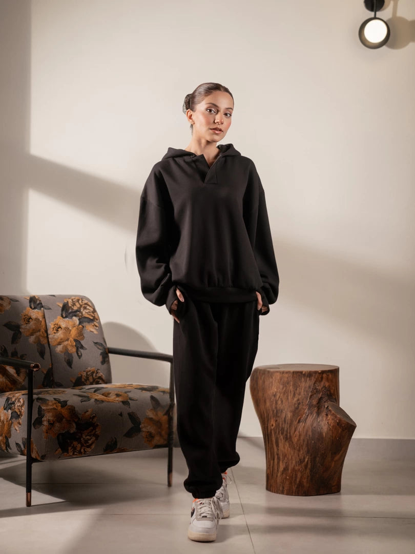 Full-length studio photograph of the Garconne oversized black hoodie, styled with black trousers and white sneakers.