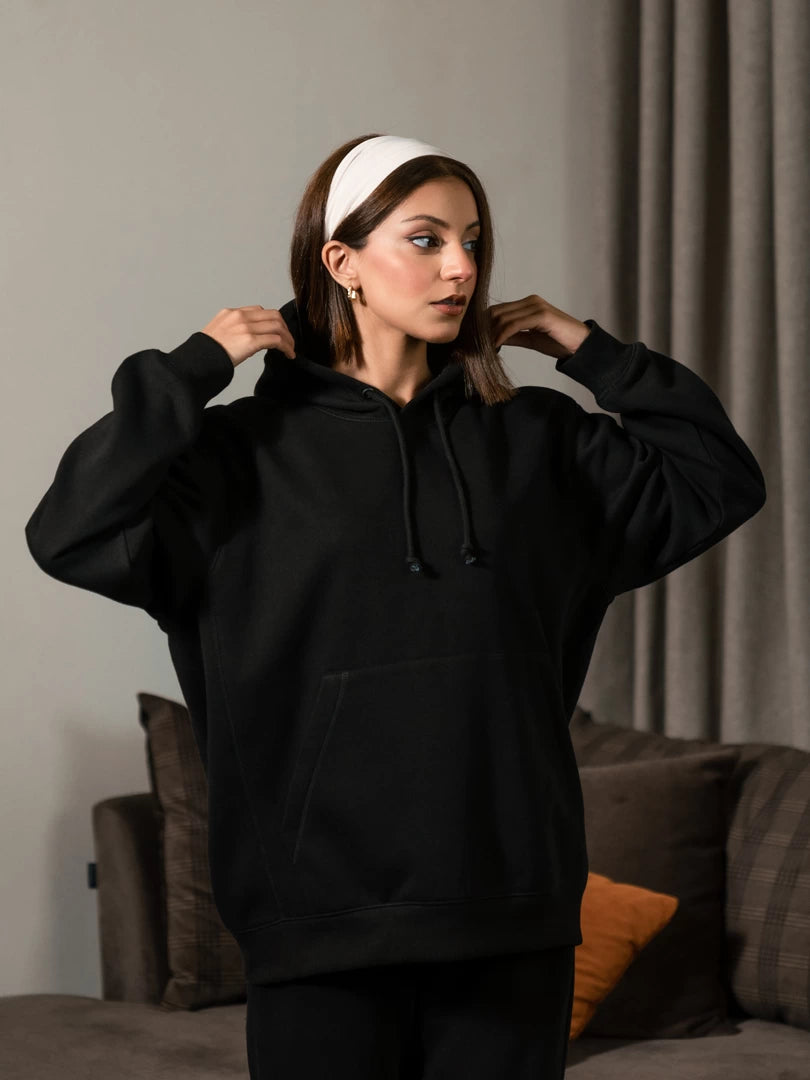 Model wearing the Lynn Essential oversized hoodie in Black, holding the hood near her shoulders to show its relaxed shape and long sleeves.