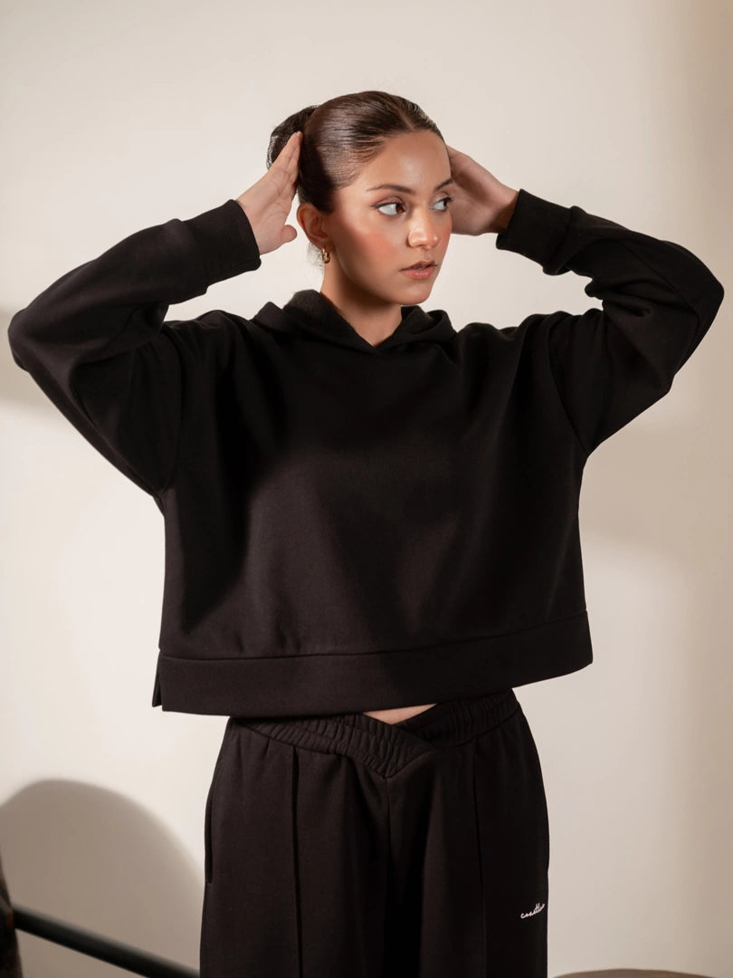 Model wearing the Soho black hoodie with both hands raised near her head, showing the boxy shape and ribbed sleeve cuffs.